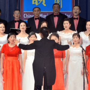 A choir group in red and black attire performing on stage under a sign that reads "china music inc." with a conductor leading them.